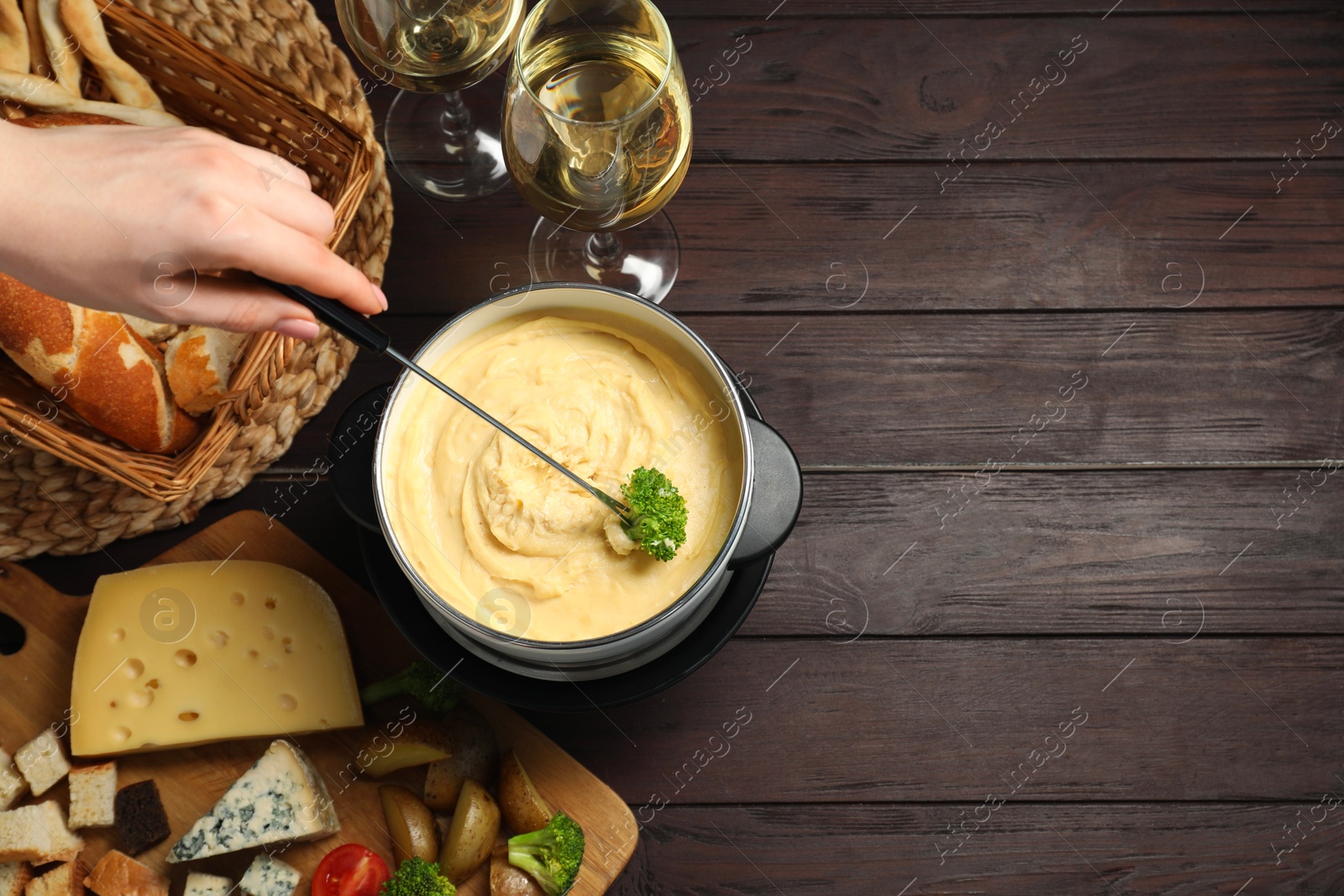 Photo of Woman dipping piece of broccoli into fondue pot with melted cheese at wooden table with wine and snacks, top view. Space for text