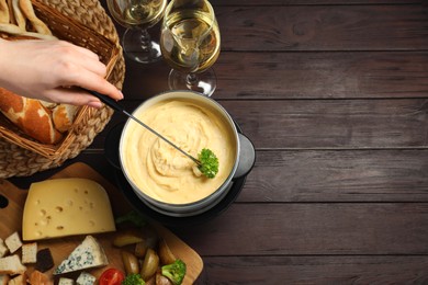 Woman dipping piece of broccoli into fondue pot with melted cheese at wooden table with wine and snacks, top view. Space for text