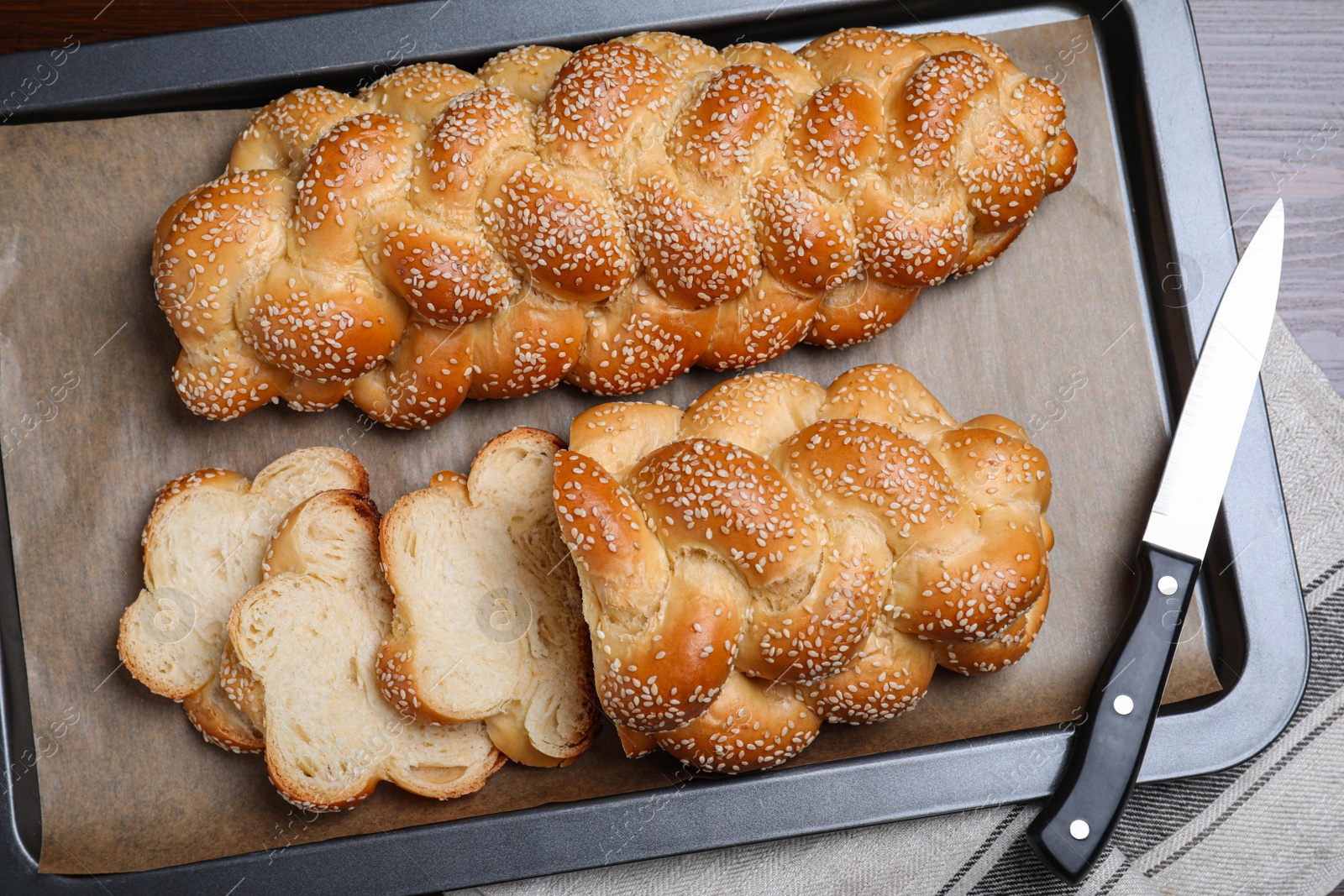 Photo of Baking tray with homemade braided bread and knife on wooden table, top view. Traditional Shabbat challah