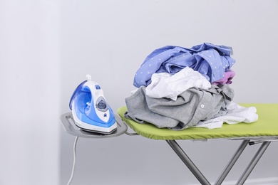 Photo of Modern electric iron and heap of clothes on board indoors. Laundry day