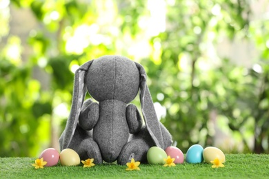 Photo of Cute Easter bunny toy and dyed eggs on green grass against blurred background, space for text