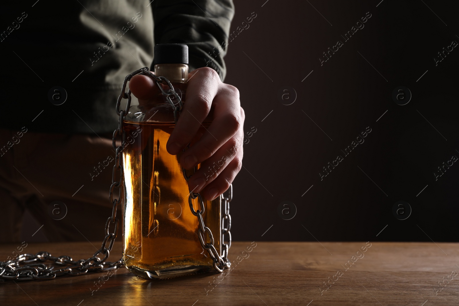 Photo of Alcohol addiction. Man chained with bottle of whiskey at wooden table, closeup. Space for text