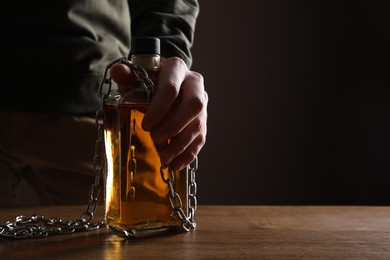Photo of Alcohol addiction. Man chained with bottle of whiskey at wooden table, closeup. Space for text
