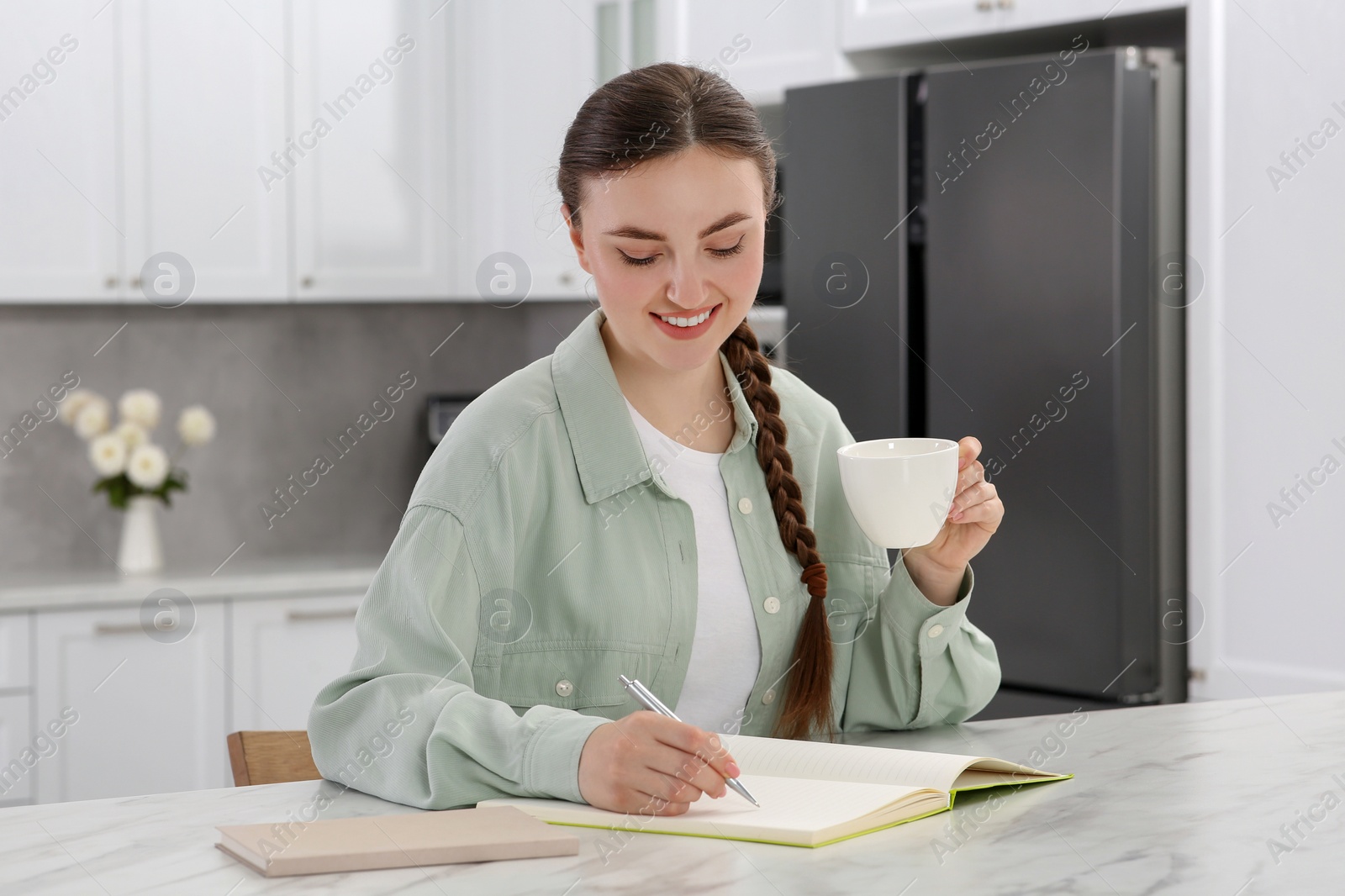 Photo of Happy young woman with cup of coffee writing in notebook at white marble table indoors