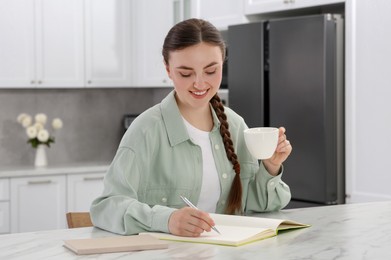 Happy young woman with cup of coffee writing in notebook at white marble table indoors