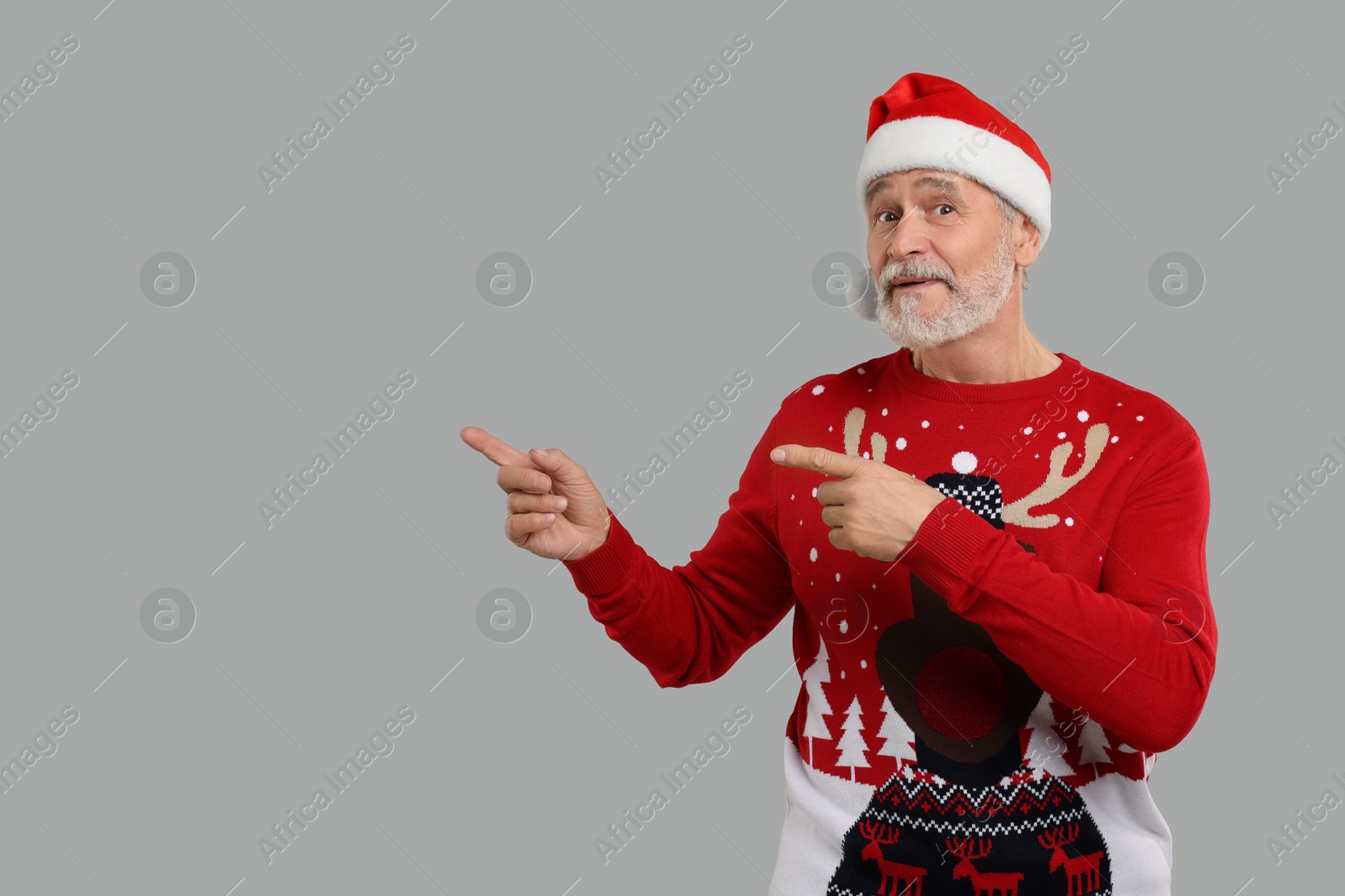 Photo of Senior man in Christmas sweater and Santa hat pointing at something on grey background. Space for text