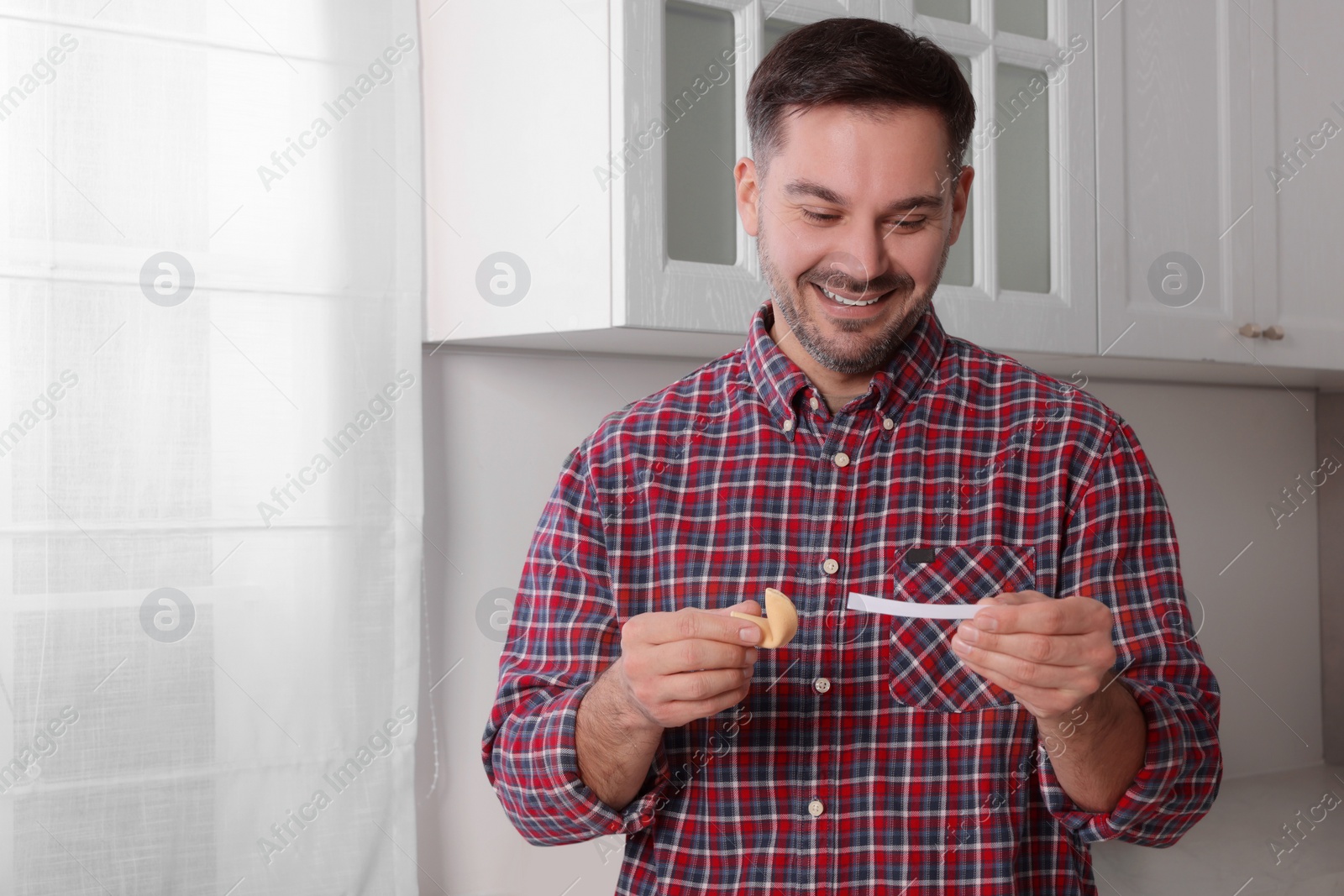 Photo of Happy man holding tasty fortune cookie and reading prediction indoors. Space for text