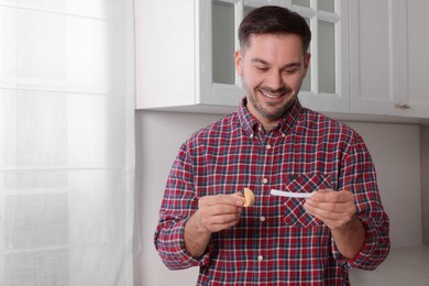 Photo of Happy man holding tasty fortune cookie and reading prediction indoors. Space for text