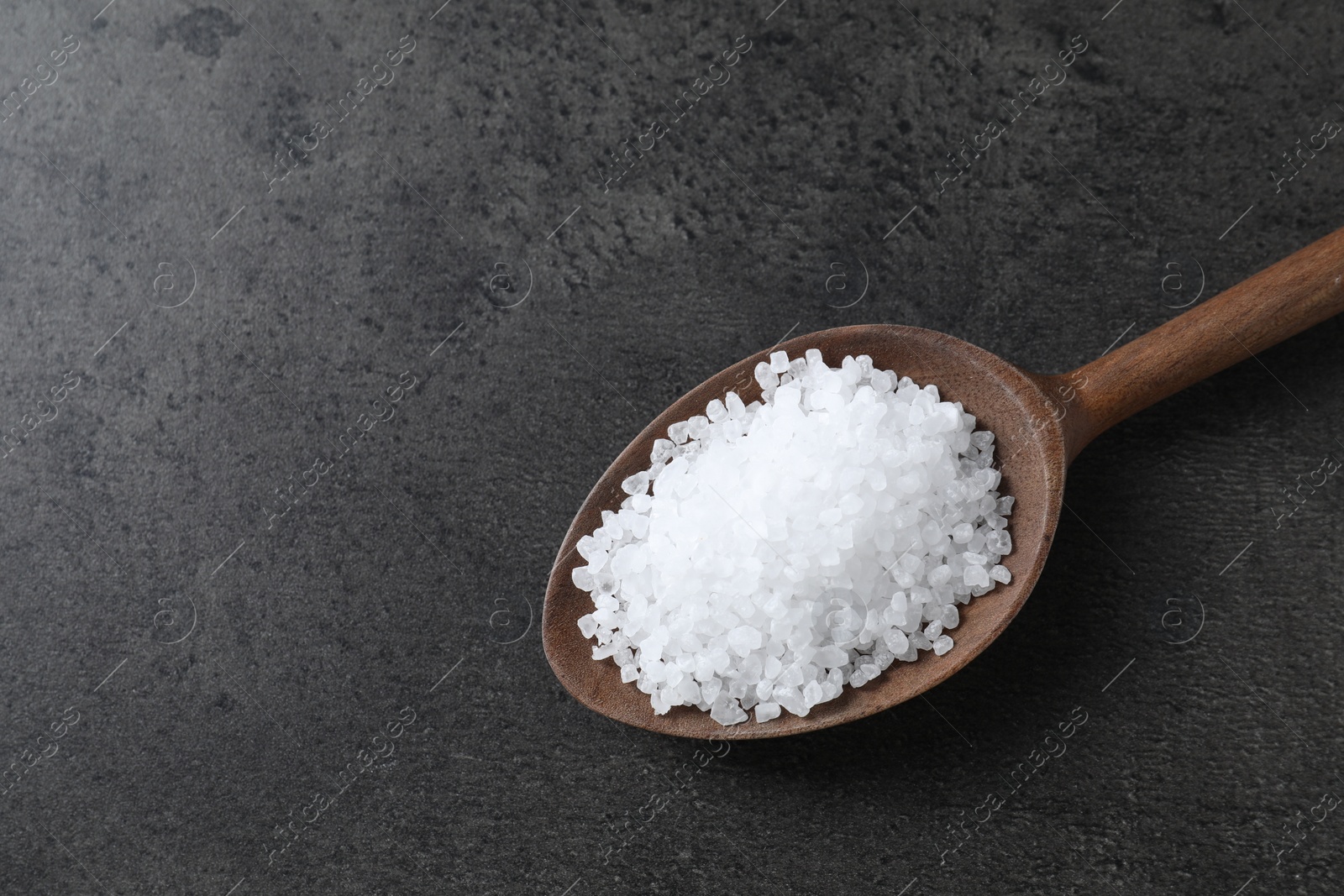 Photo of Natural salt in wooden spoon on dark grey table, top view. Space for text