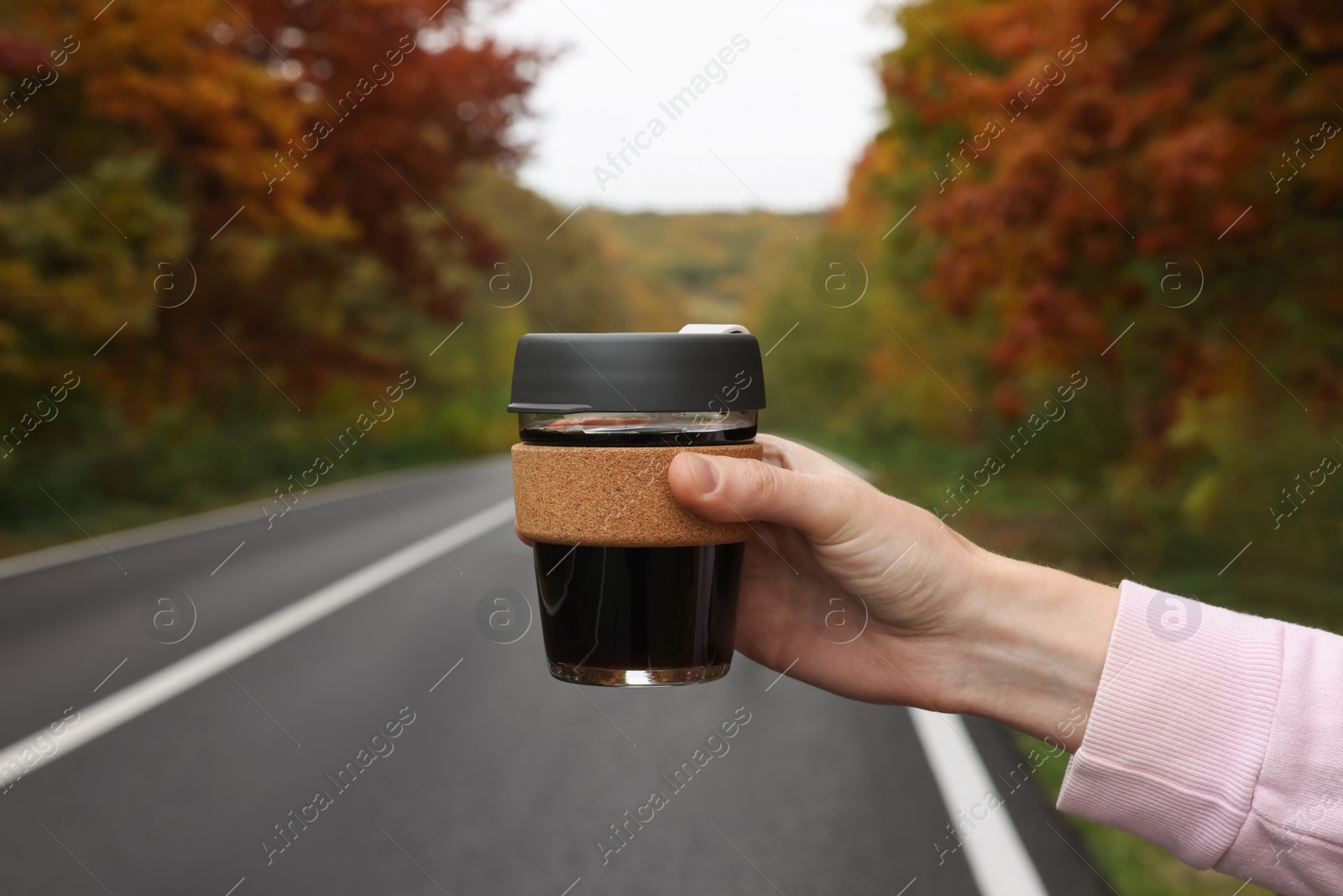 Photo of Woman holding reusable glass cup with hot drink near road outdoors, closeup. Autumn season
