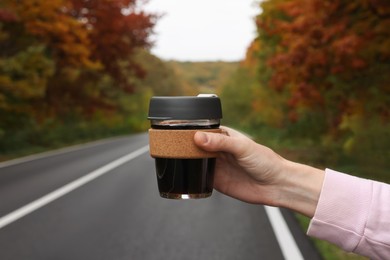 Woman holding reusable glass cup with hot drink near road outdoors, closeup. Autumn season