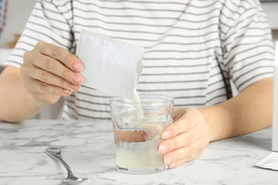 Photo of Woman pouring powder from medicine sachet into glass with water at table, closeup