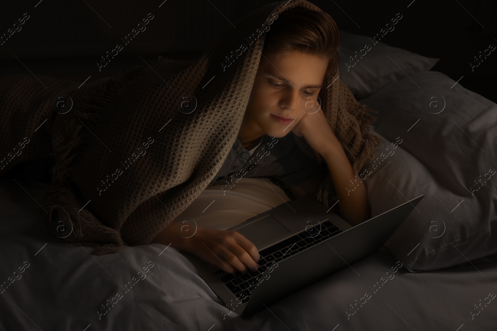 Photo of Teenage boy using laptop under blanket on bed at night. Internet addiction