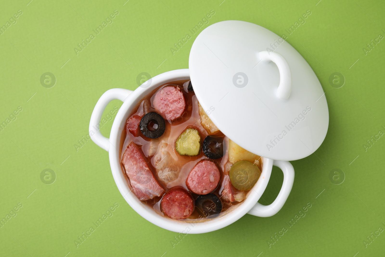 Photo of Meat solyanka soup with thin dry smoked sausages in pot on light green background, top view