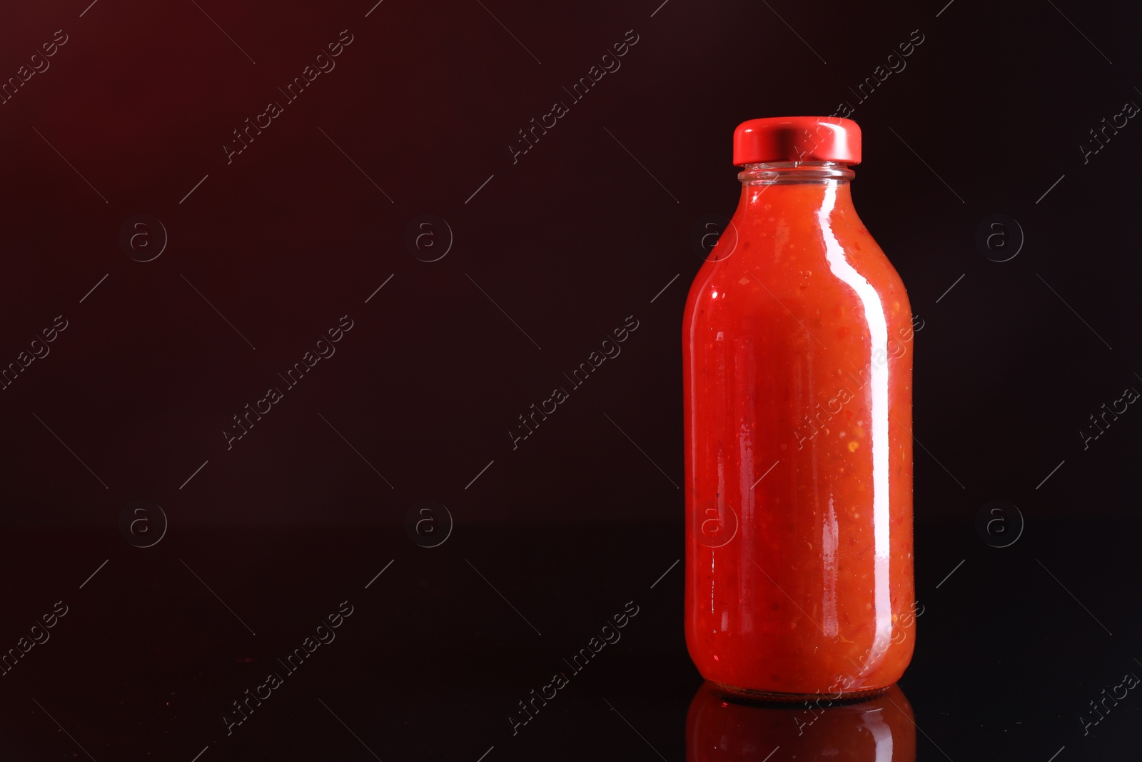 Photo of Spicy chili sauce in bottle against dark background, space for text