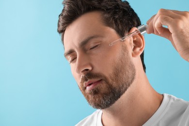 Handsome man applying cosmetic serum onto his face on light blue background, closeup. Space for text
