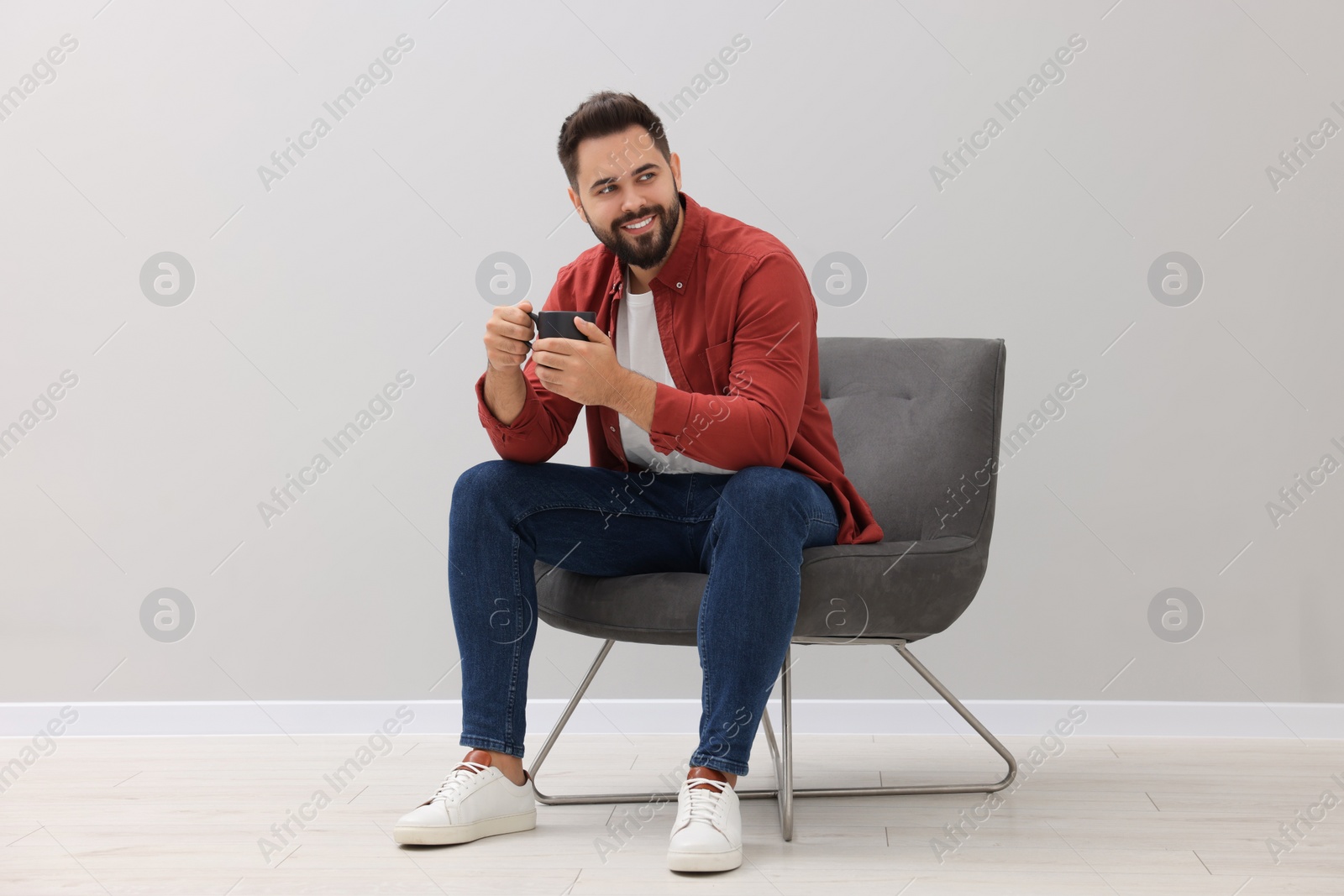 Photo of Handsome man with cup of drink sitting in armchair near light grey wall indoors