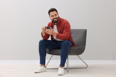 Handsome man with cup of drink sitting in armchair near light grey wall indoors