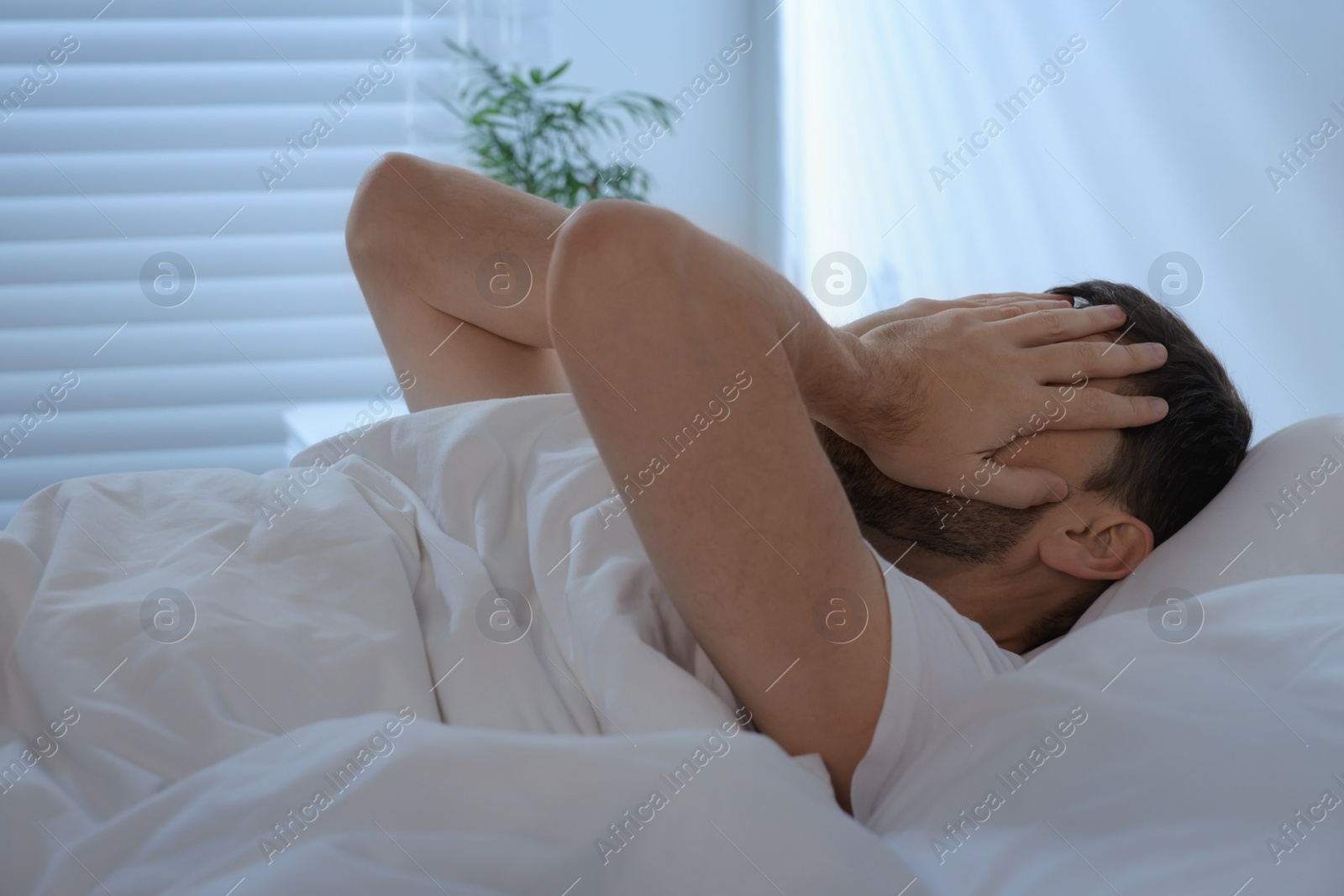 Photo of Sleepy man covering his face with hands in bed at home