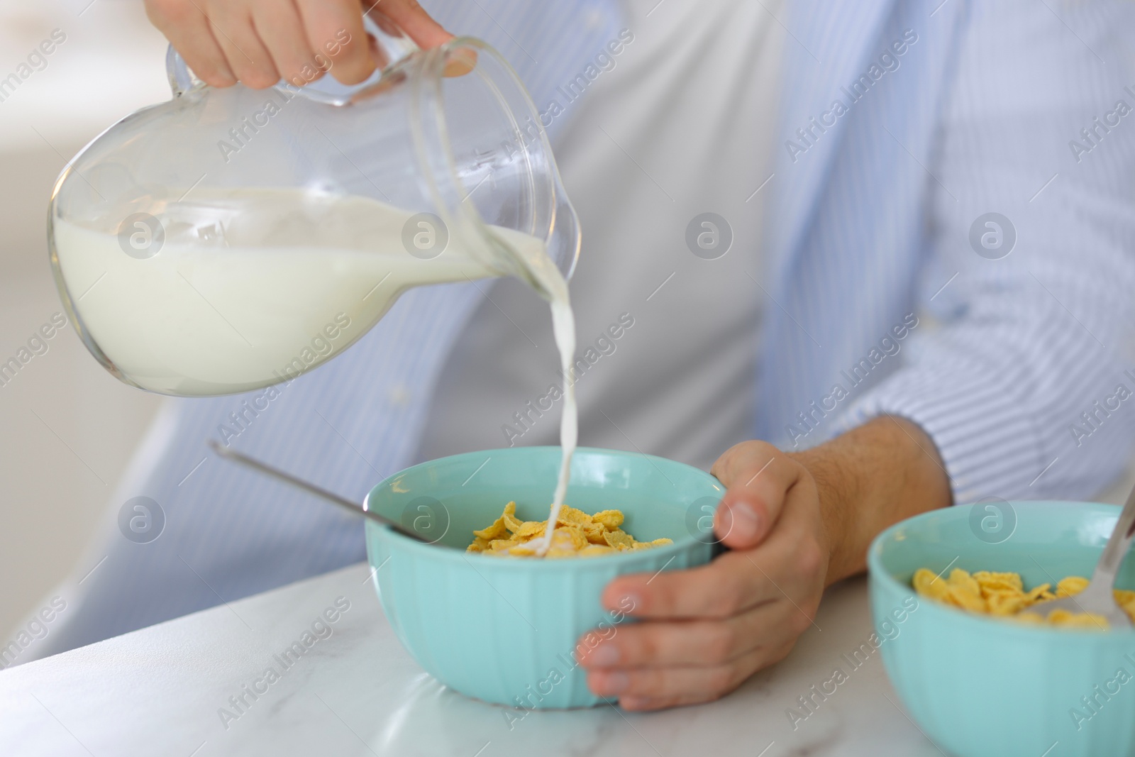 Photo of Making breakfast. Man pouring milk from jug into bowl with cornflakes at white marble table, closeup