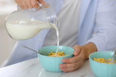 Photo of Making breakfast. Man pouring milk from jug into bowl with cornflakes at white marble table, closeup