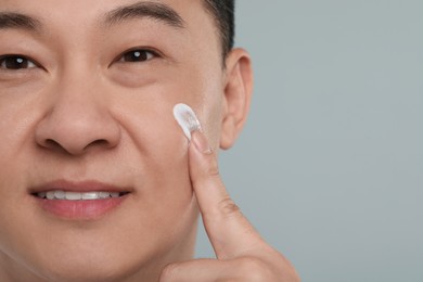 Handsome man applying cream onto his face on light grey background, closeup. Space for text