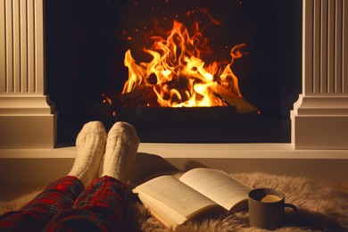 Photo of Woman with cup of drink and book near fireplace at home, closeup