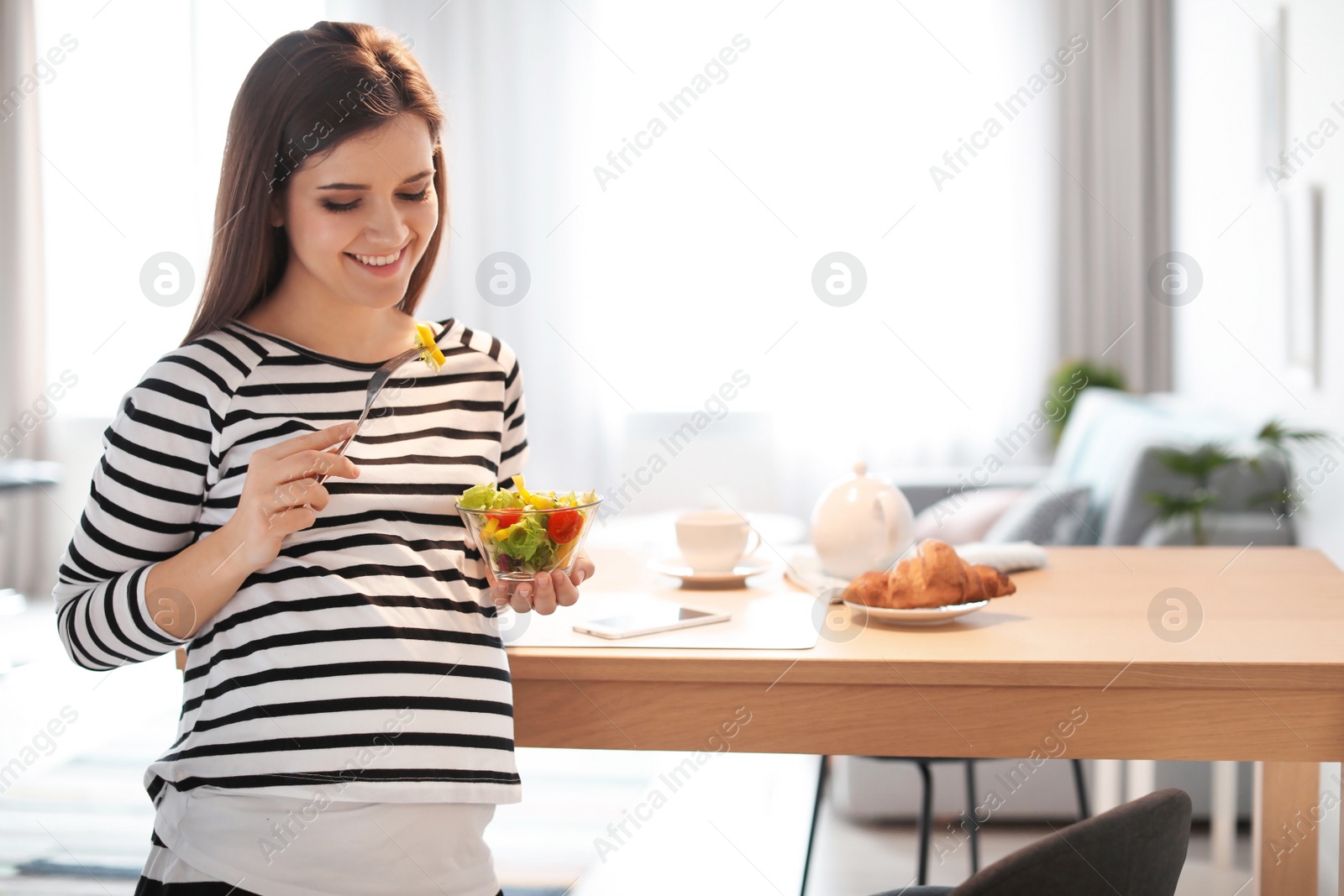 Photo of Young pregnant woman eating vegetable salad near table in kitchen