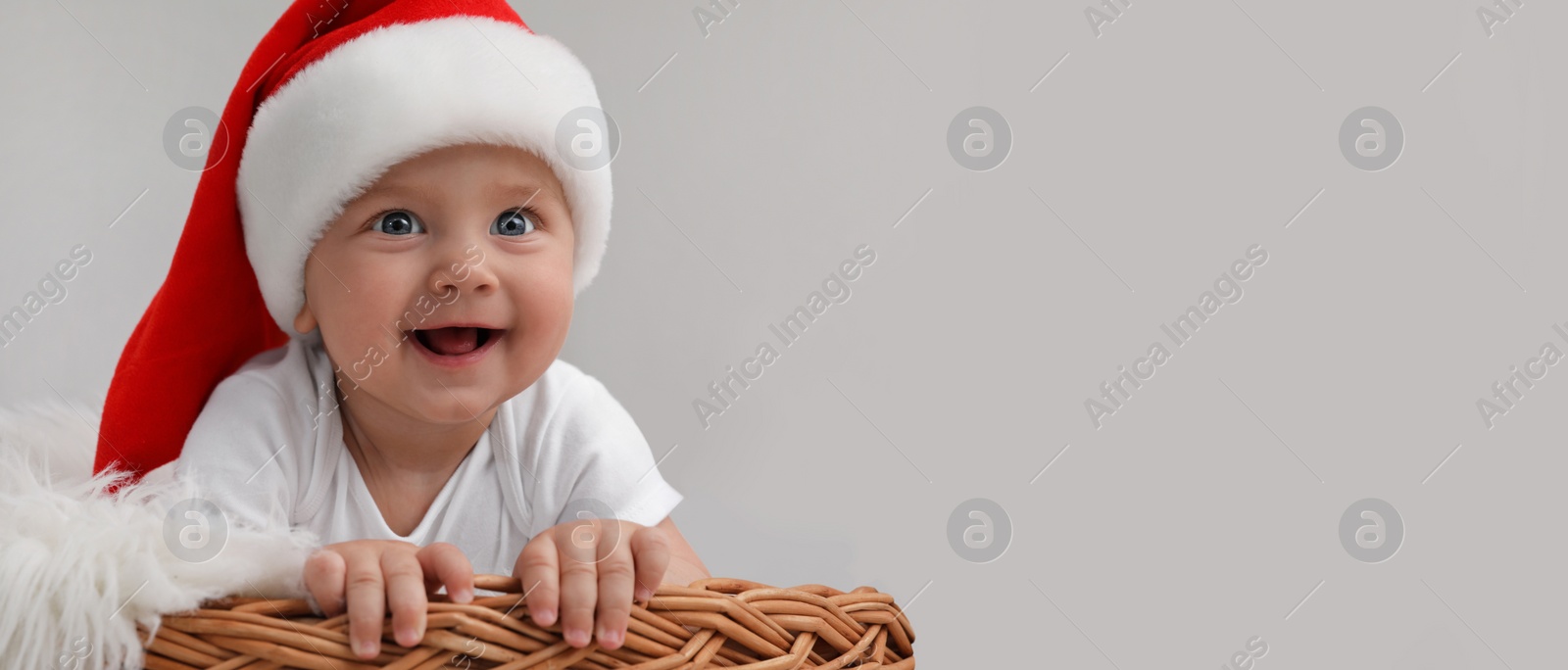 Image of Cute baby wearing Santa hat in wicker basket on light grey background, banner design with space for text. Christmas celebration