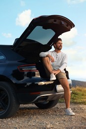 Photo of Happy man sitting in trunk of modern car on roadside outdoors