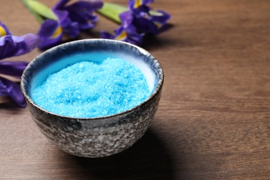 Photo of Light blue sea salt in bowl and flowers on wooden table, closeup. Space for text