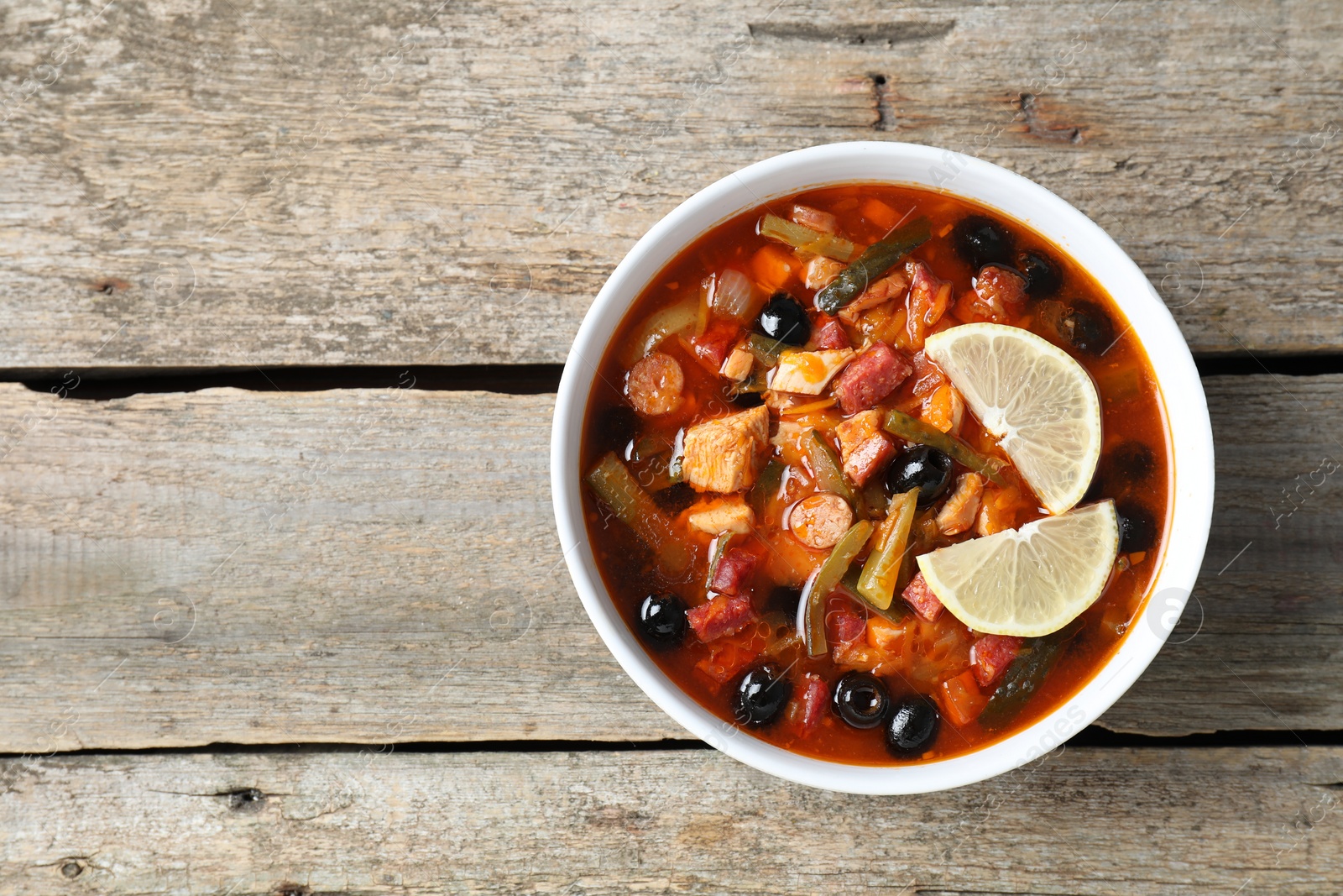 Photo of Meat solyanka soup with sausages, olives and vegetables in bowl on wooden table, top view. Space for text