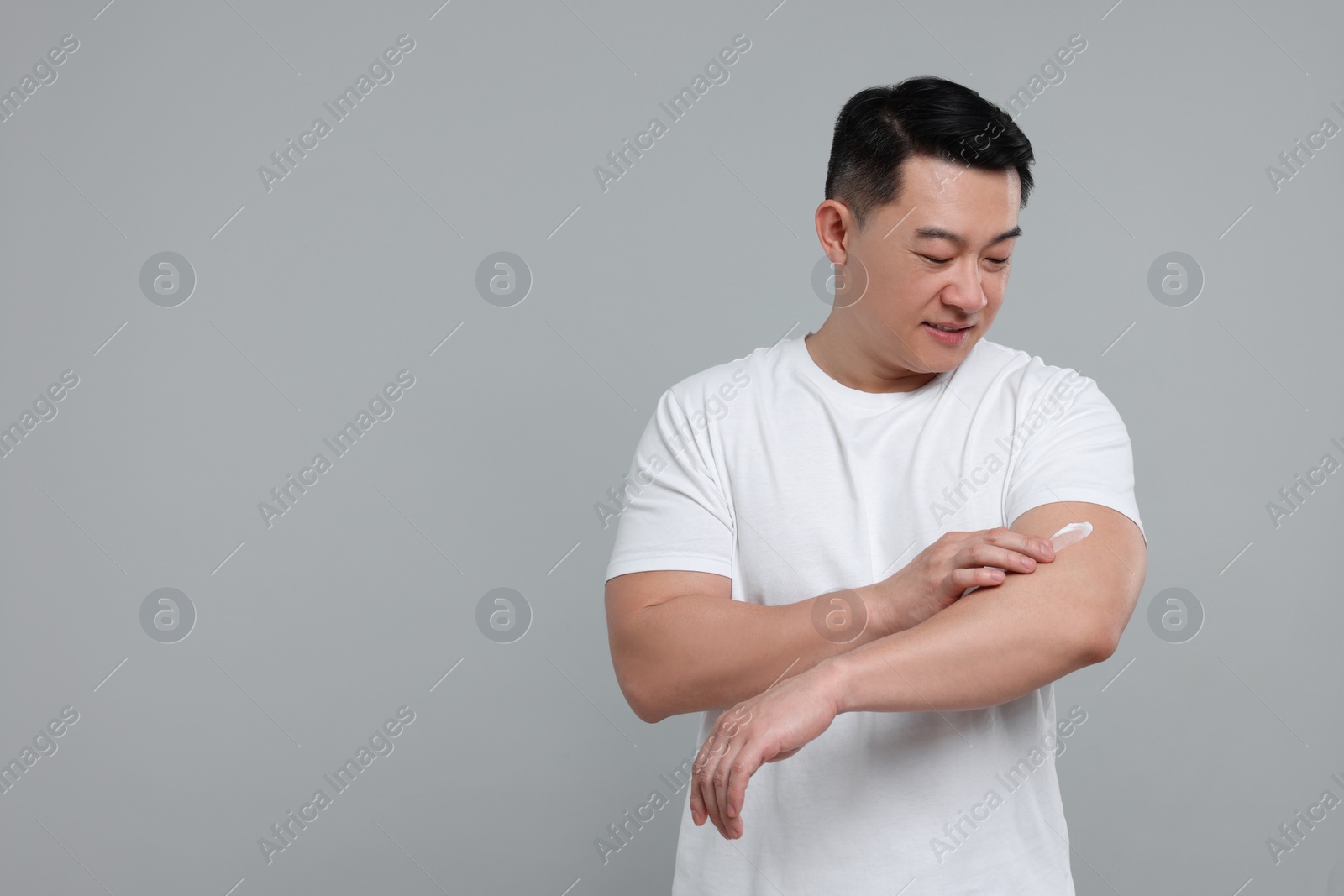 Photo of Handsome man applying body cream onto his arm on light grey background. Space for text