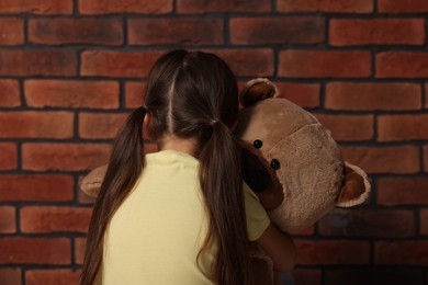 Photo of Child abuse. Upset little girl with teddy bear near brick wall, back view