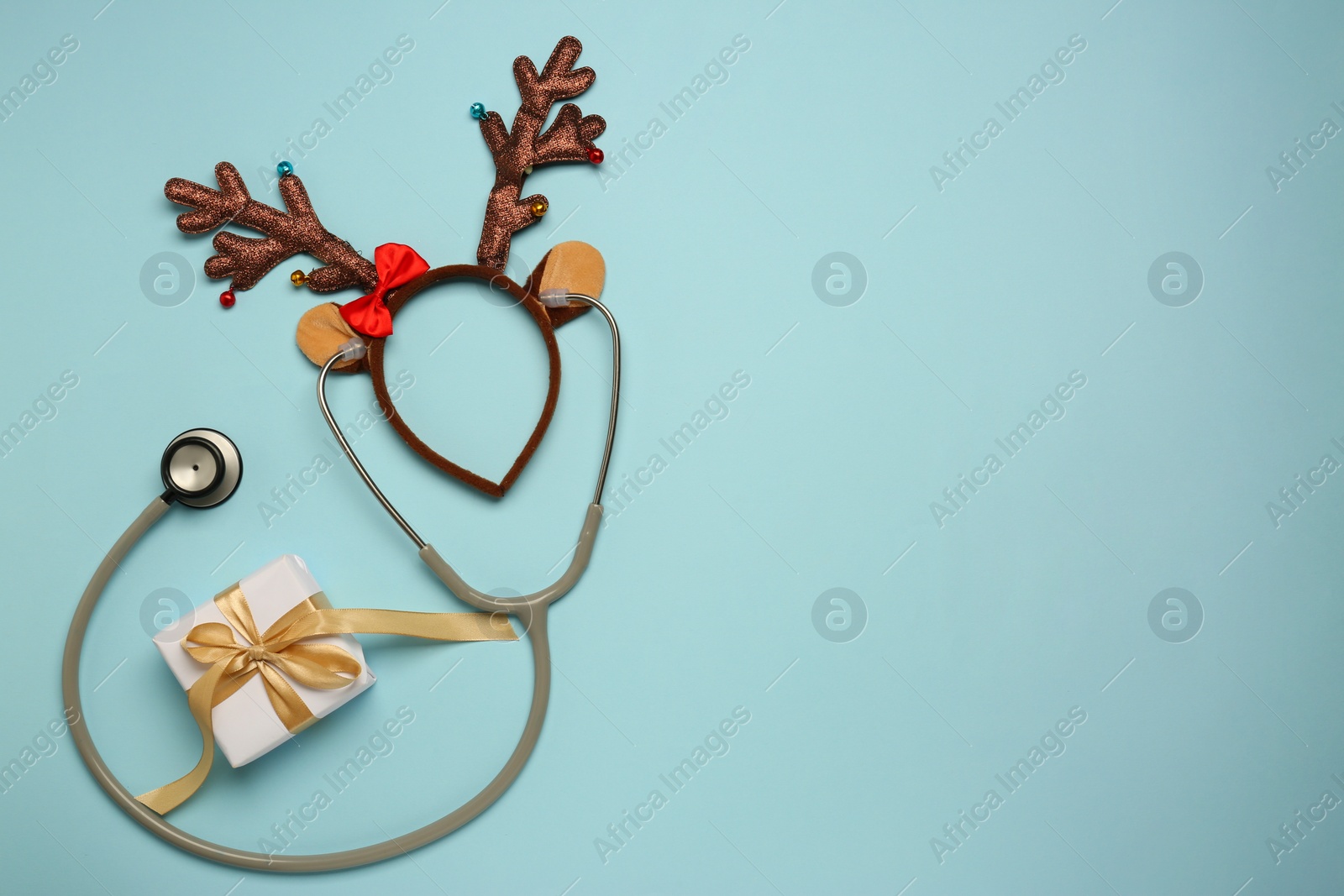 Photo of Greeting card for doctor with stethoscope, gift box and reindeer headband on light blue background, flat lay. Space for text