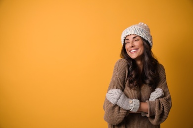 Young woman in warm sweater, hat and mittens on yellow background, space for text. Christmas season