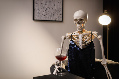 Skeleton in dress with wine sitting at table indoors