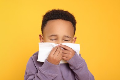 Photo of African-American boy blowing nose in tissue on yellow background. Cold symptoms