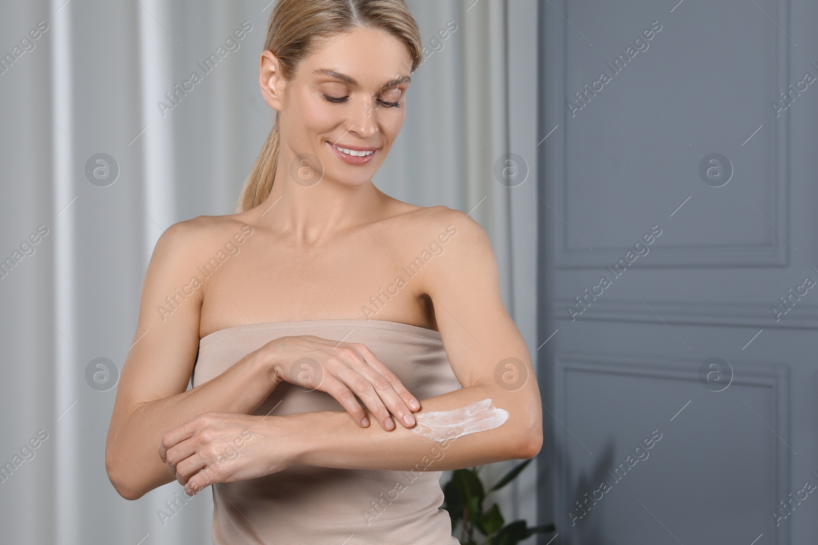 Photo of Happy woman applying body cream onto arm indoors, space for text