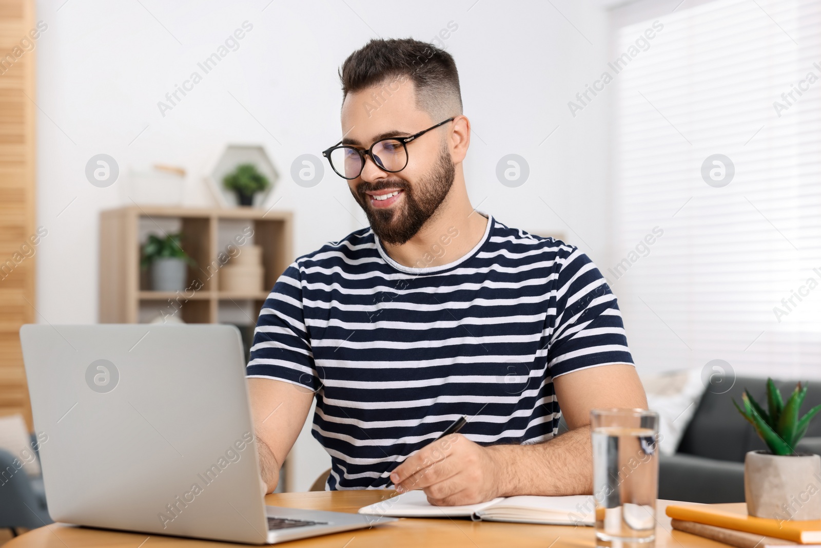 Photo of Young man writing in notebook while working on laptop at wooden table indoors