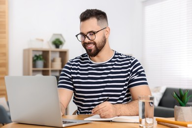 Photo of Young man writing in notebook while working on laptop at wooden table indoors