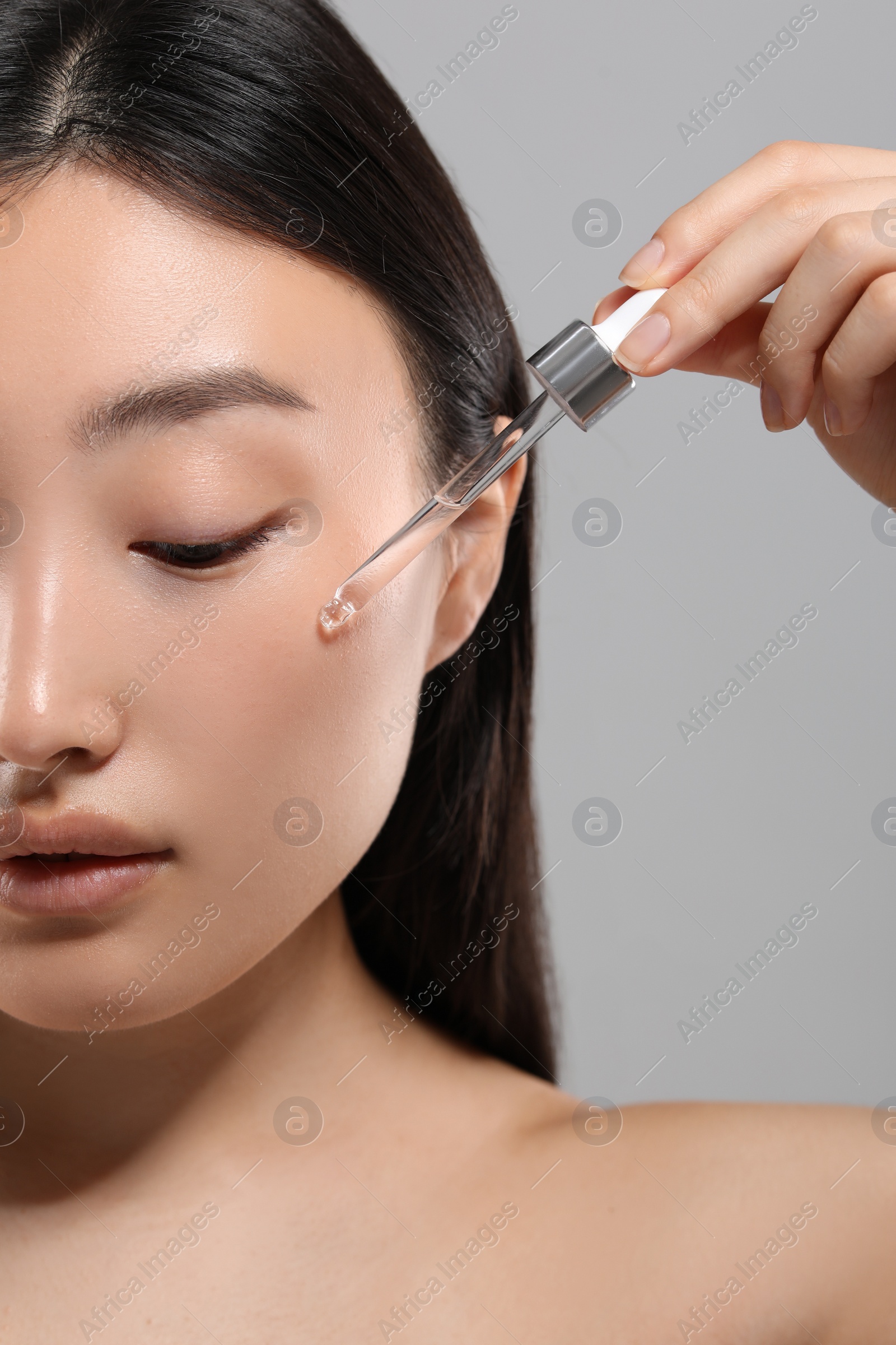 Photo of Beautiful young woman applying cosmetic serum onto her face on grey background, closeup