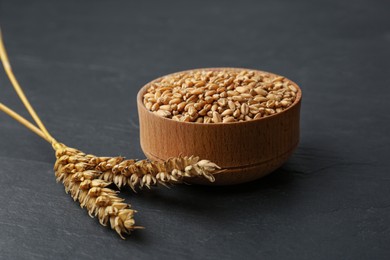 Photo of Dried ears of wheat and grains in wooden bowl on black table