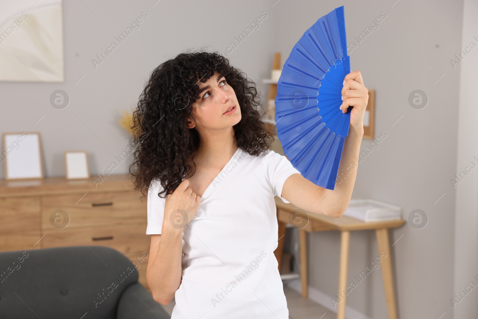 Photo of Young woman waving blue hand fan to cool herself at home