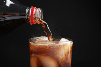 Photo of Pouring refreshing cola from bottle into glass with ice cubes on black background, closeup