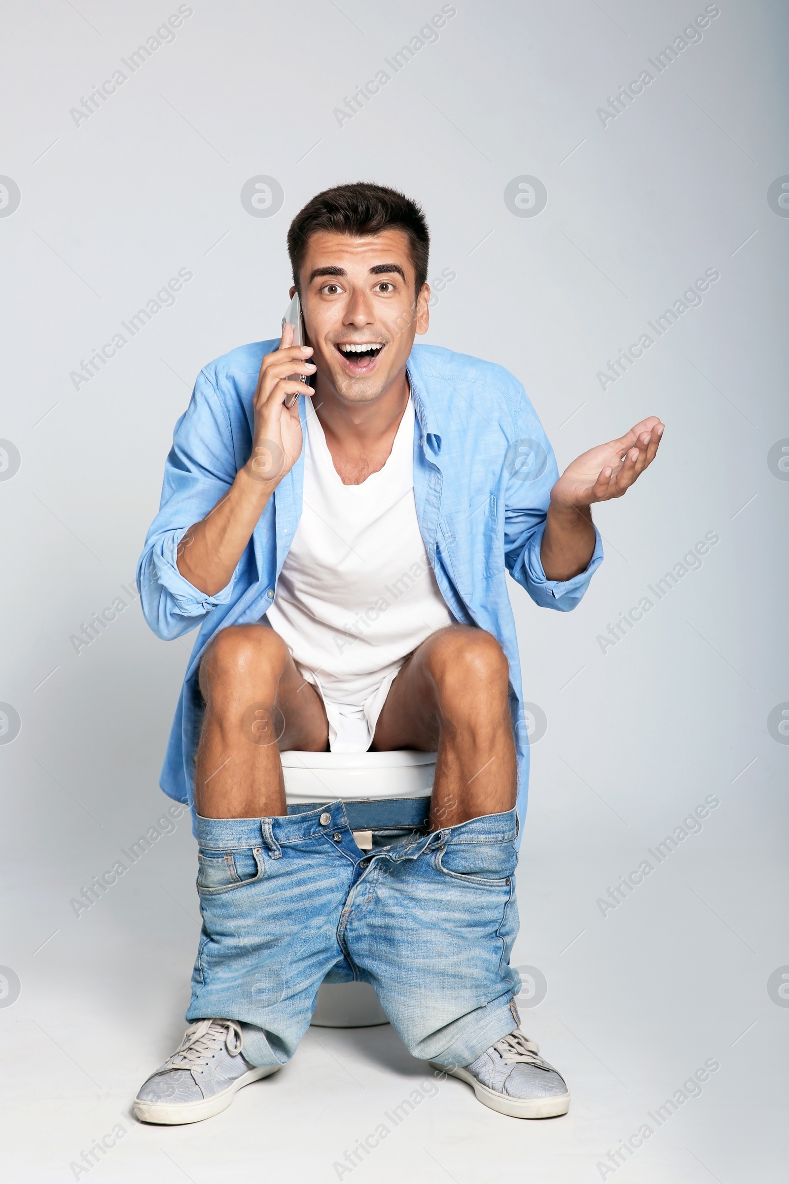 Photo of Young man with mobile phone sitting on toilet bowl against gray background