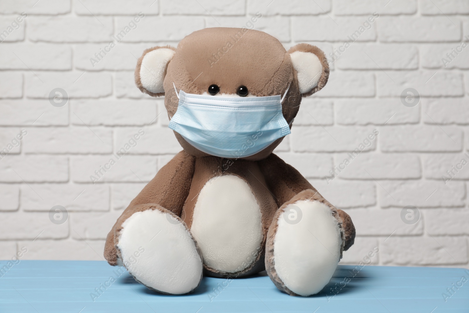 Photo of Cute teddy bear with medical mask on light blue wooden table near white brick wall