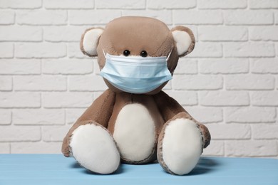 Photo of Cute teddy bear with medical mask on light blue wooden table near white brick wall