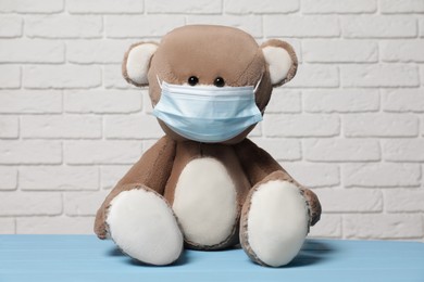 Cute teddy bear with medical mask on light blue wooden table near white brick wall