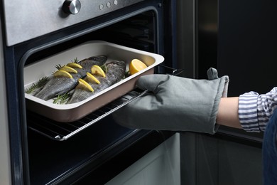 Woman putting baking tray with sea bass fish, lemon and rosemary into oven, closeup